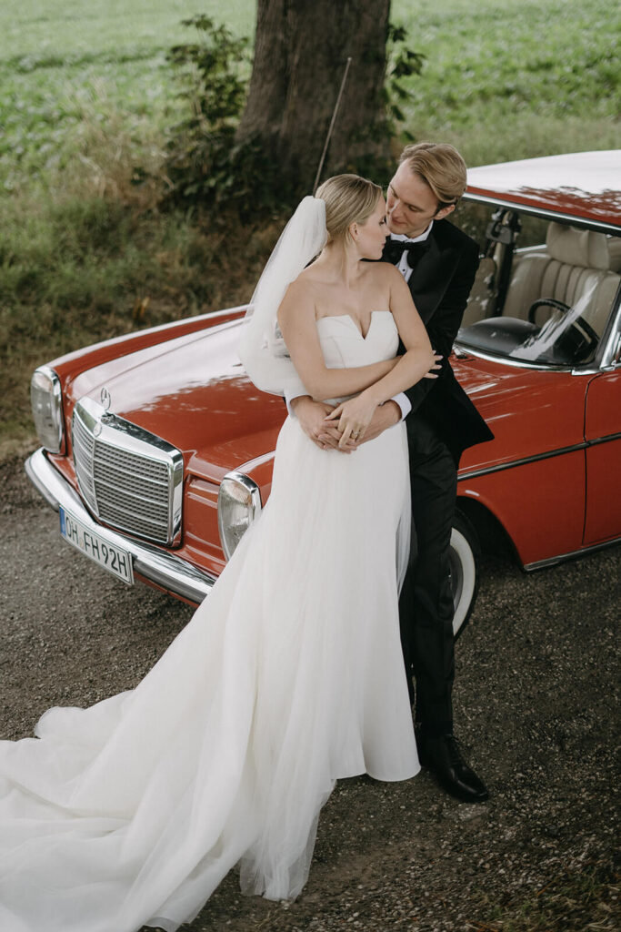 A newly married couple in suit and beautiful white minimalistic wedding dress posing in front of a red old-timer car.